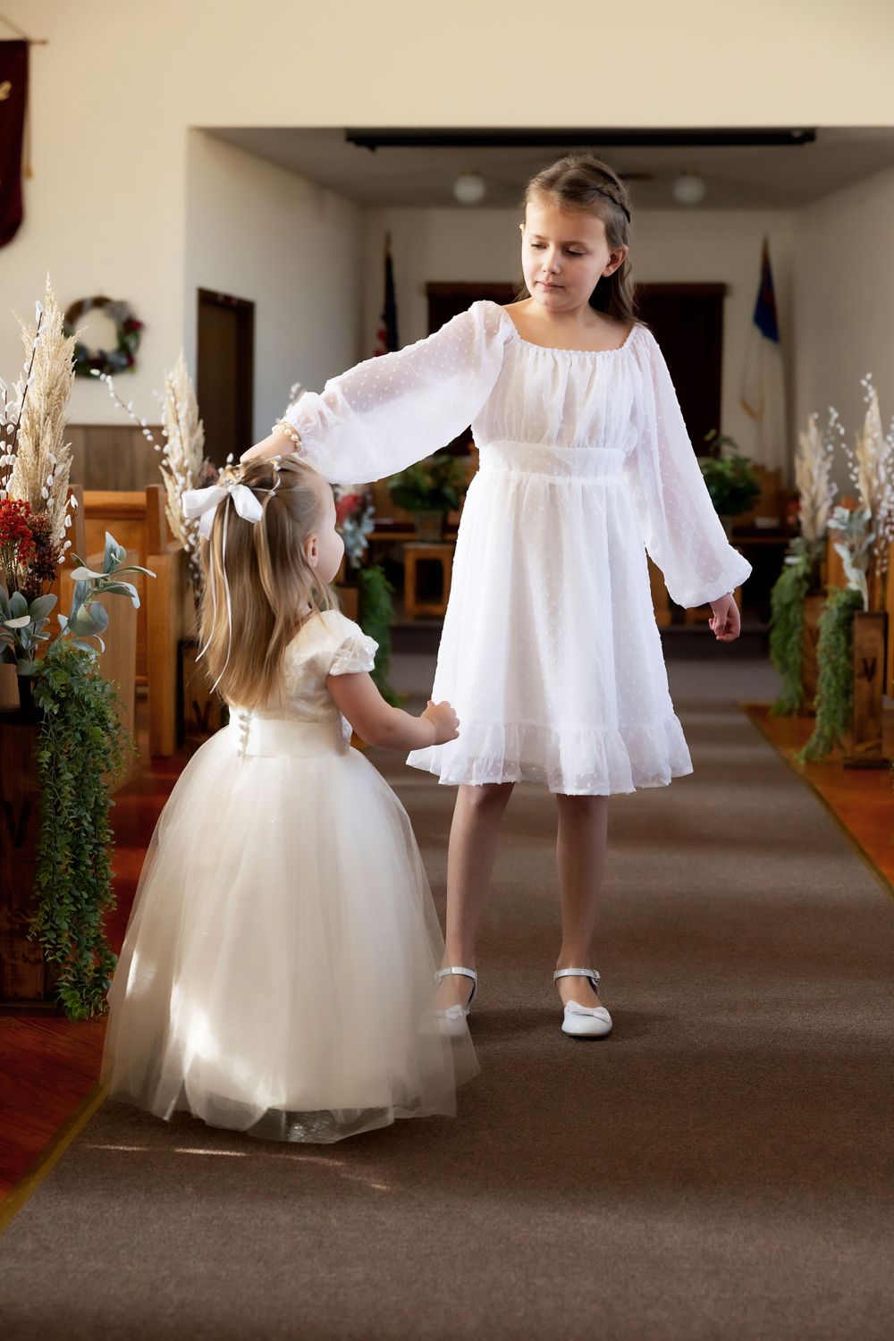 Young attendants in white dresses share a sweet moment in church aisle decorated with flowers.