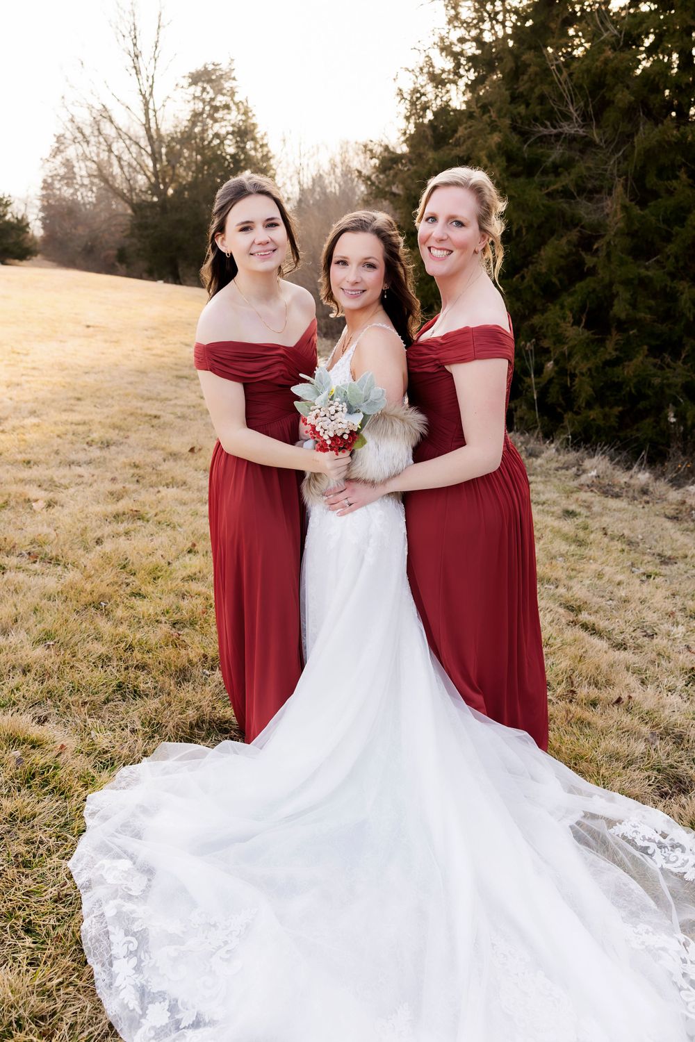 Three women in burgundy and white gowns pose together in an outdoor setting at sunset.