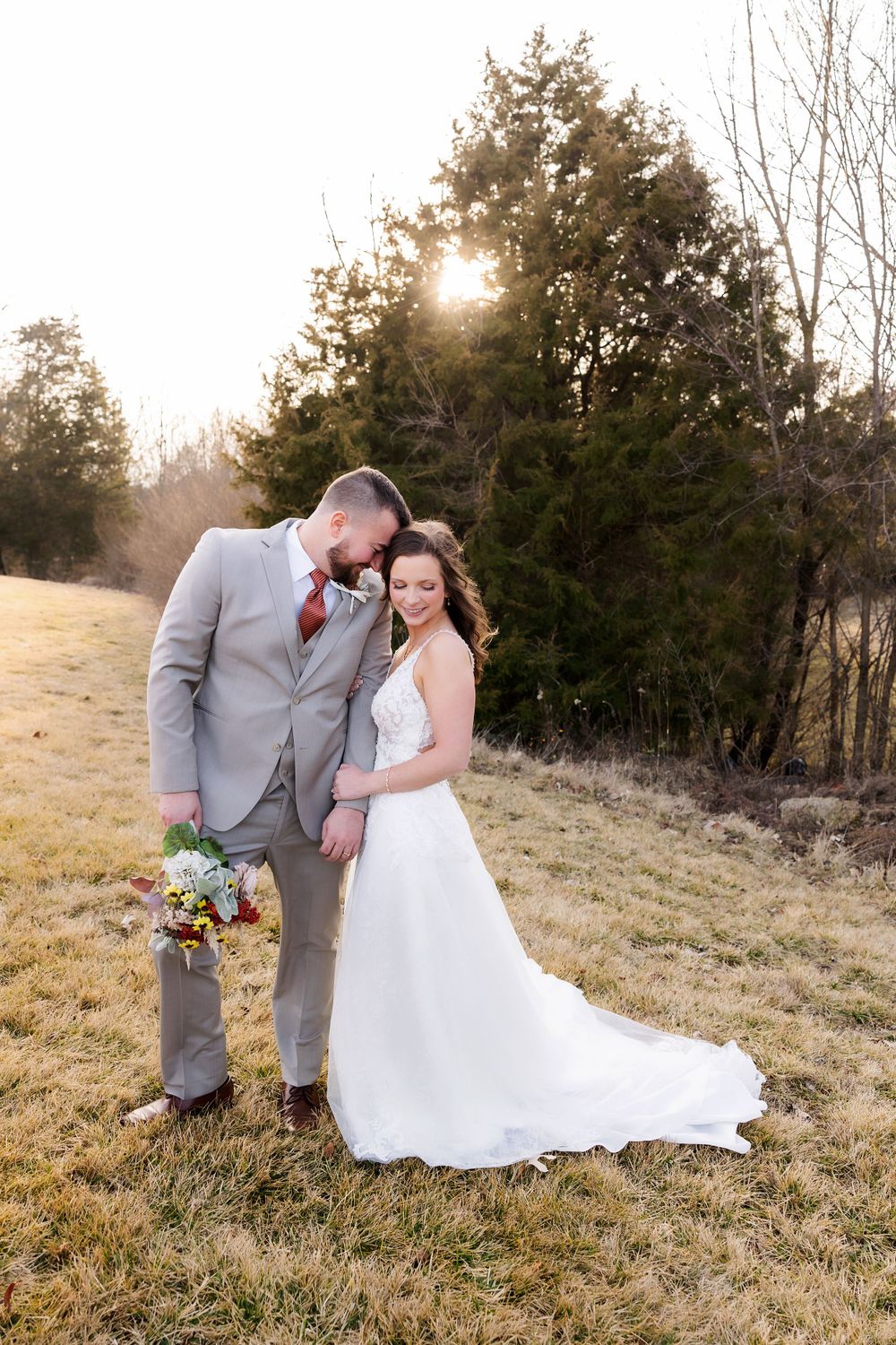 A couple in wedding attire poses together against a sunset backdrop with evergreen trees.