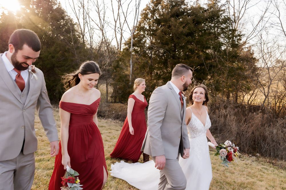 Wedding party with bridesmaids in burgundy dresses walks through a sunlit field.