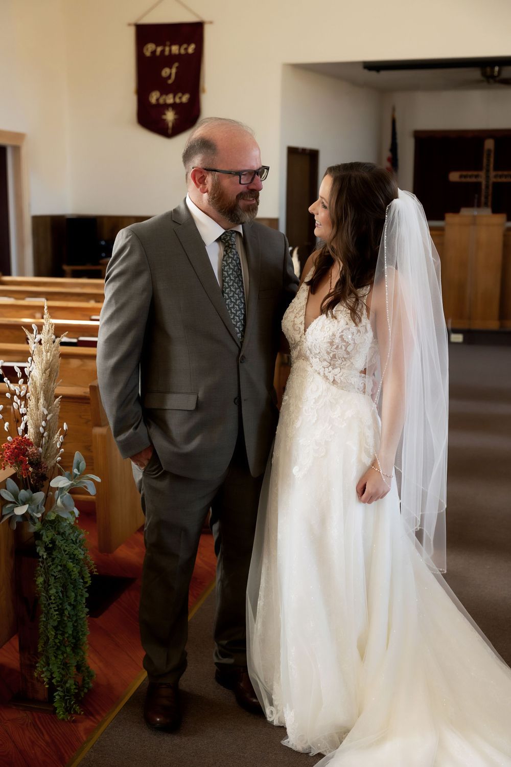 A touching moment between a bride in a lace gown and suited companion inside a church with pews and banners.