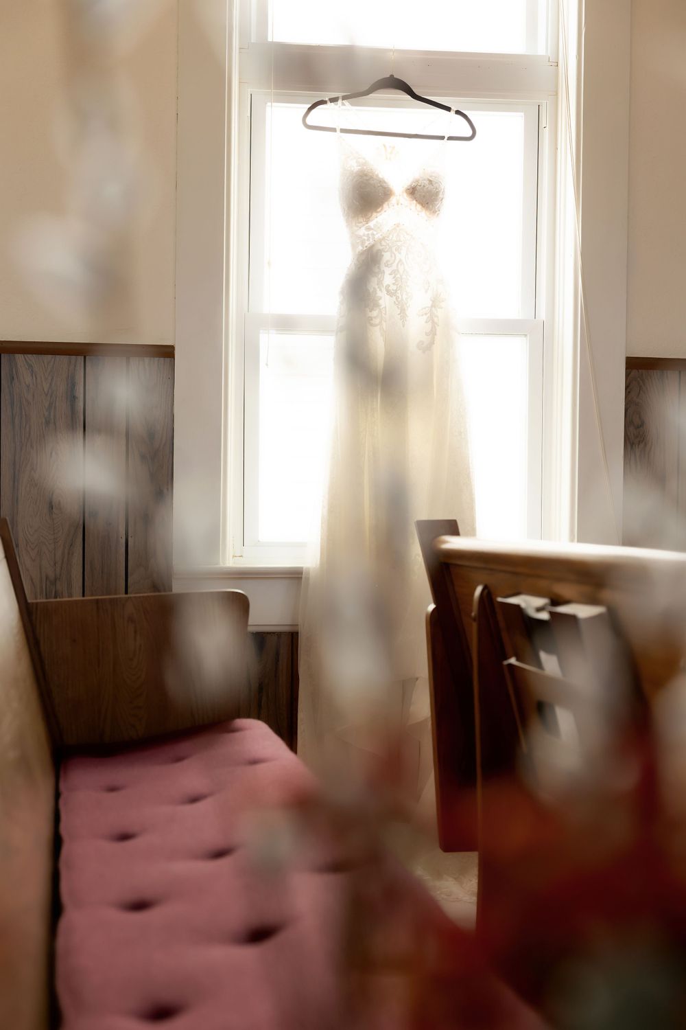 White wedding dress hanging in window light with pink church pew visible below.