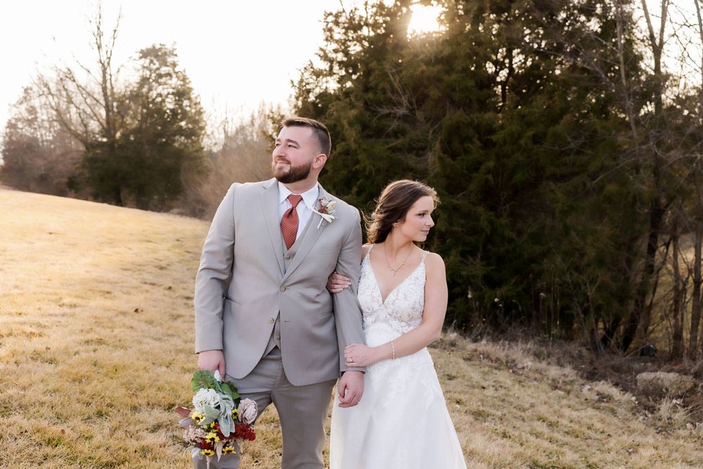 A wedding couple poses in a sunlit field with evergreen trees in the background.