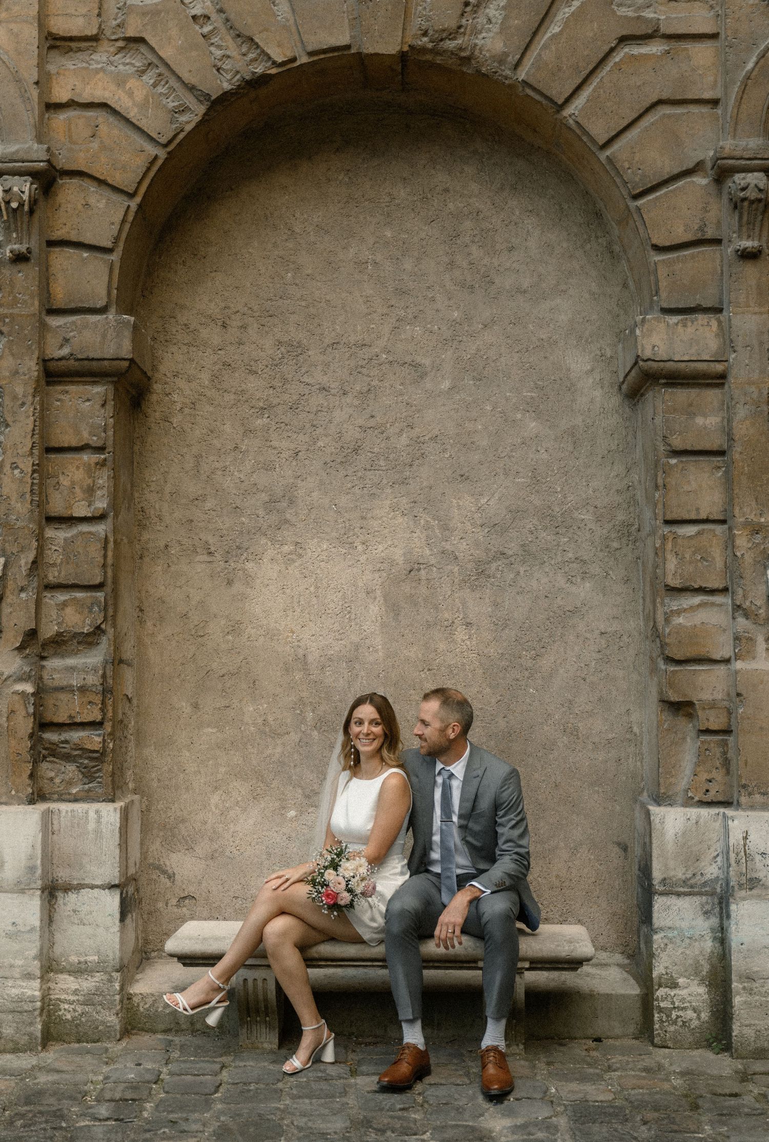 A couple sits together in an ornate stone archway alcove in elegant wedding attire.