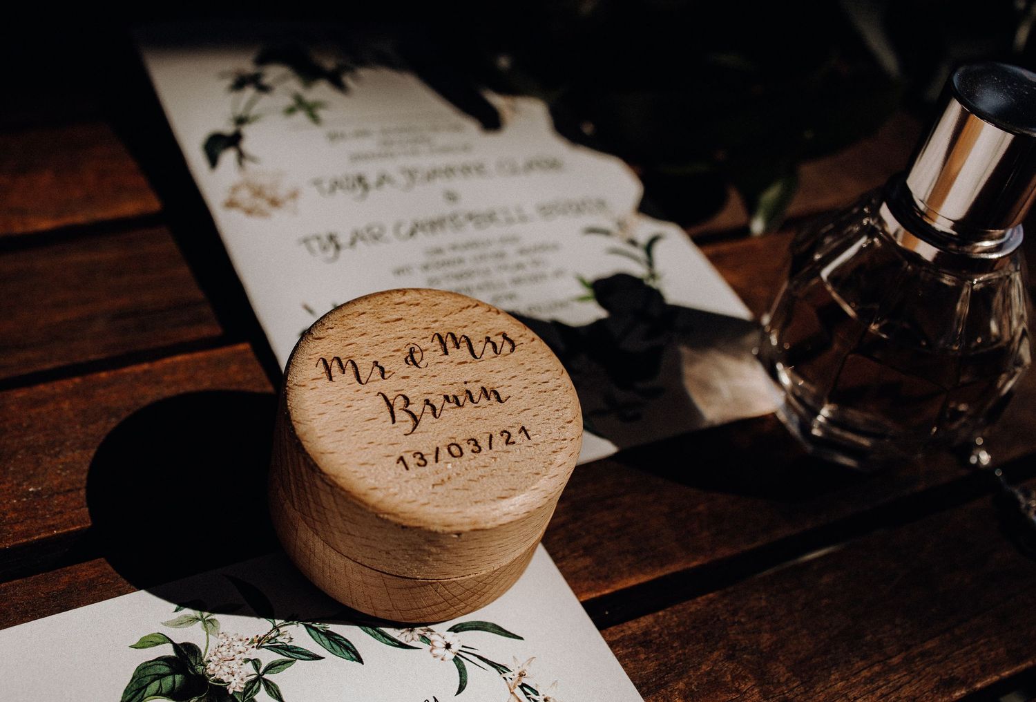Wedding invitation and wooden ring box with perfume bottle on dark wooden surface.