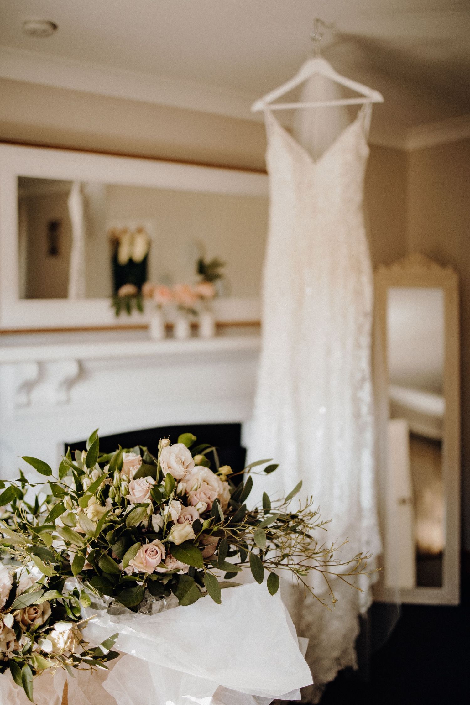 White lace wedding dress hanging above fireplace mantel with floral arrangement.