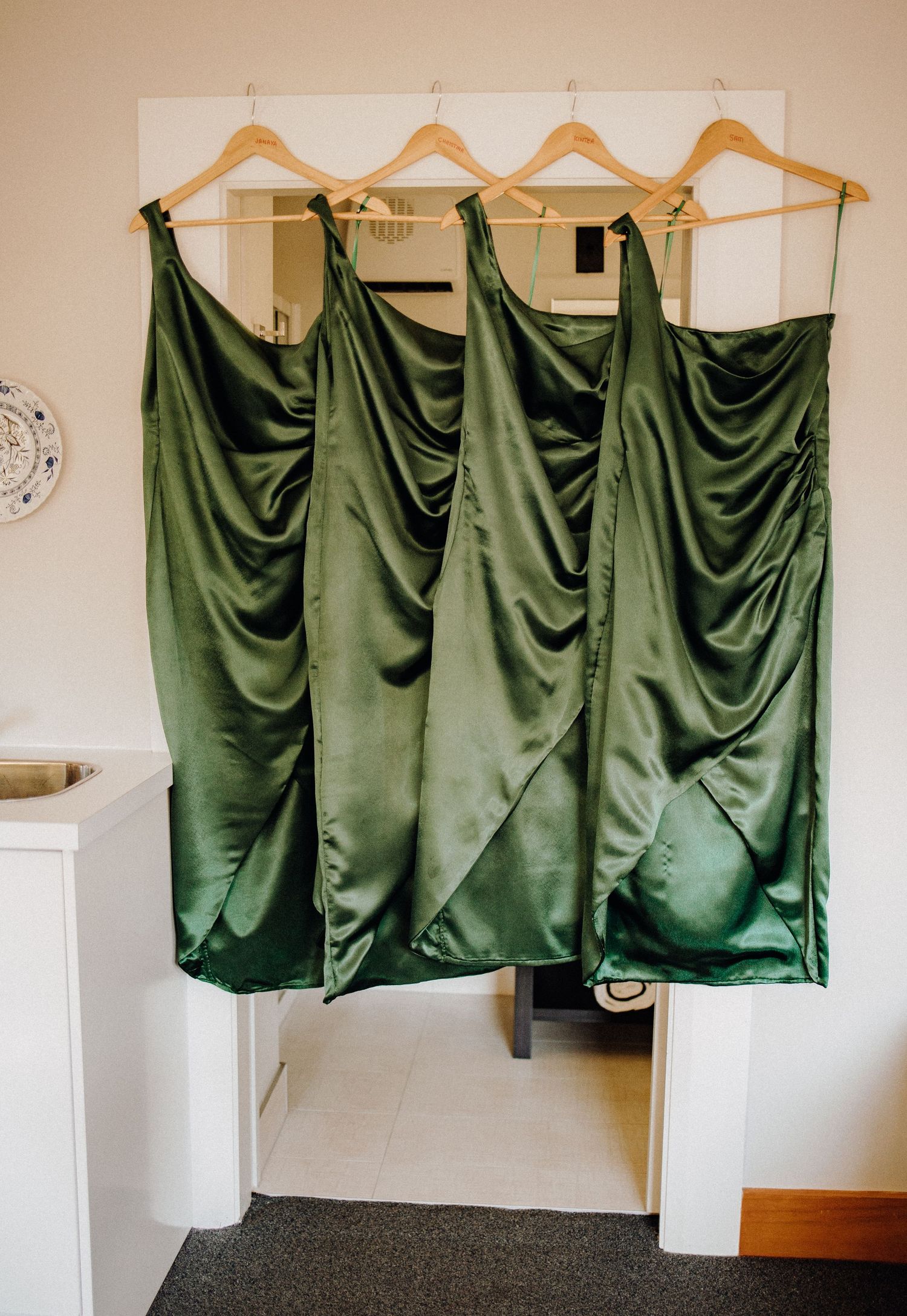 Four emerald green satin bridesmaid dresses hanging on wooden hangers against white wall.