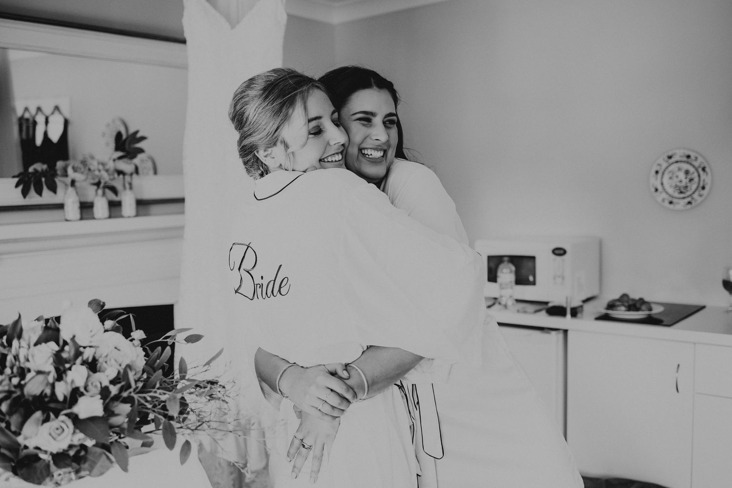 Black and white photo showing a couple embracing in a kitchen with a 'Bride' robe and flowers visible in the scene.