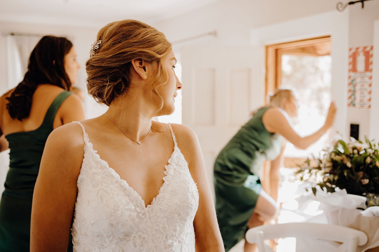 Wedding preparations in a bright room with a bride in a lace V-neck dress while others help with arrangements.