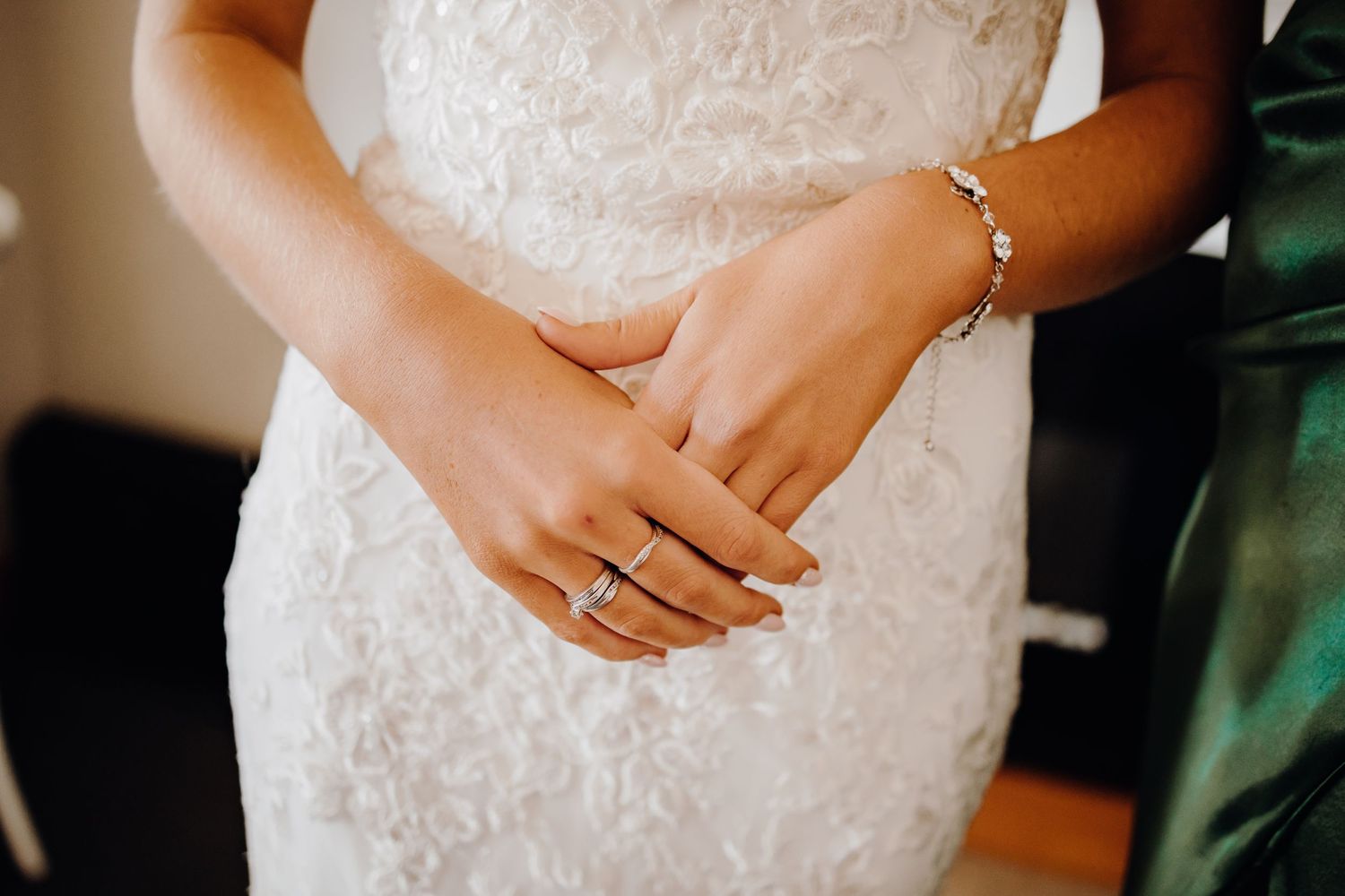 Close up detail of hands wearing wedding and engagement rings against a white lace dress.