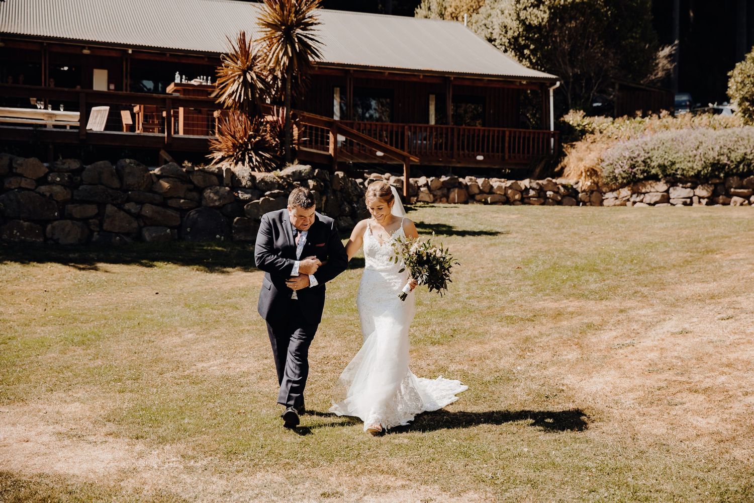 Wedding couple walks across sunny lawn towards rustic building during outdoor ceremony.