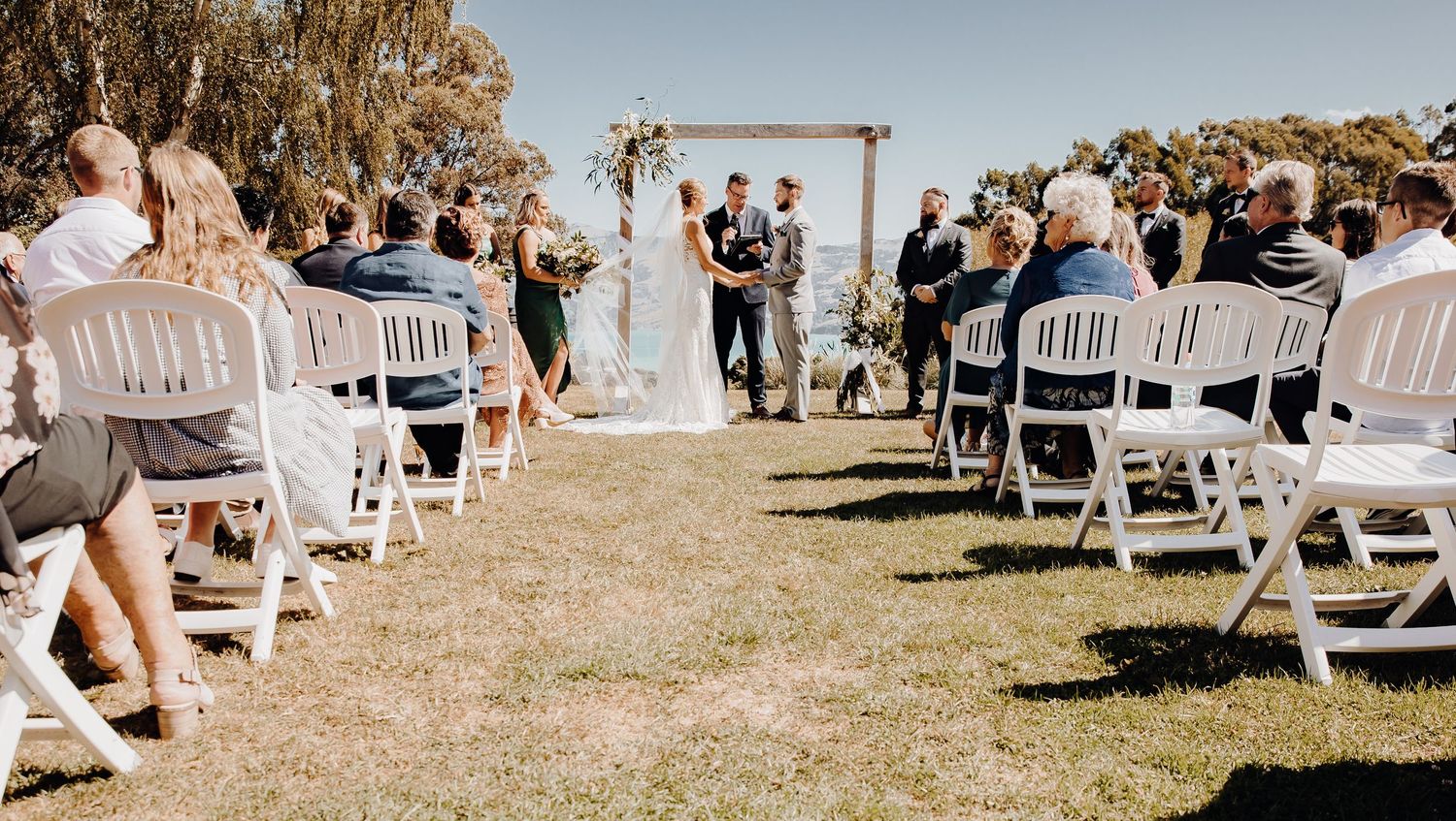 Outdoor wedding ceremony setup with white chairs and wooden arch against mountain backdrop.