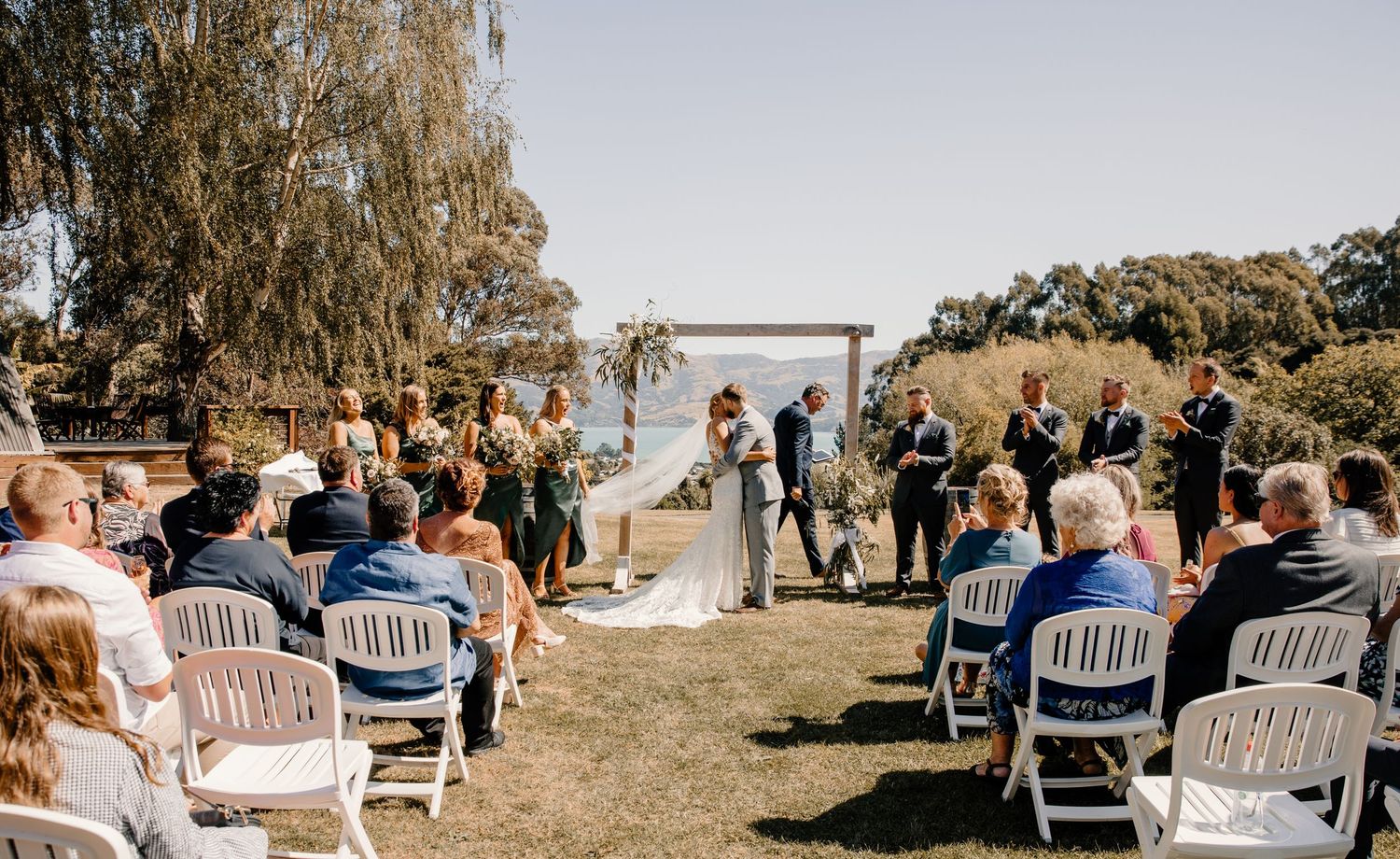 Wedding guests seated in white chairs watch ceremony under wooden arch with mountain views.