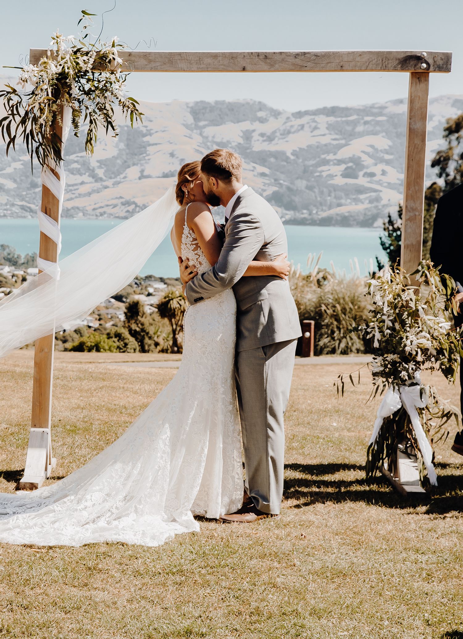 Couple shares intimate moment under floral-decorated wooden arch with lake and mountains in background.