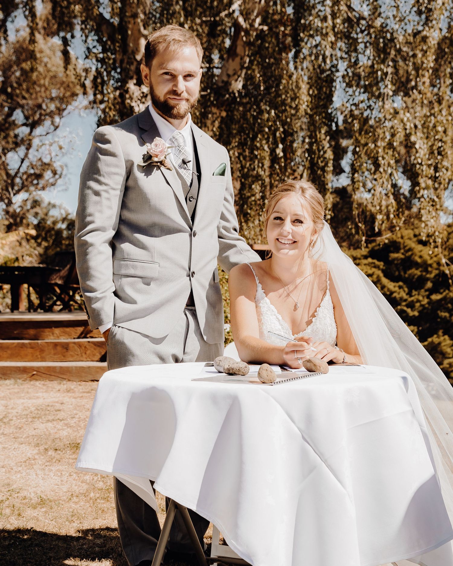 Couple signing marriage documents at white-clothed table during outdoor ceremony.