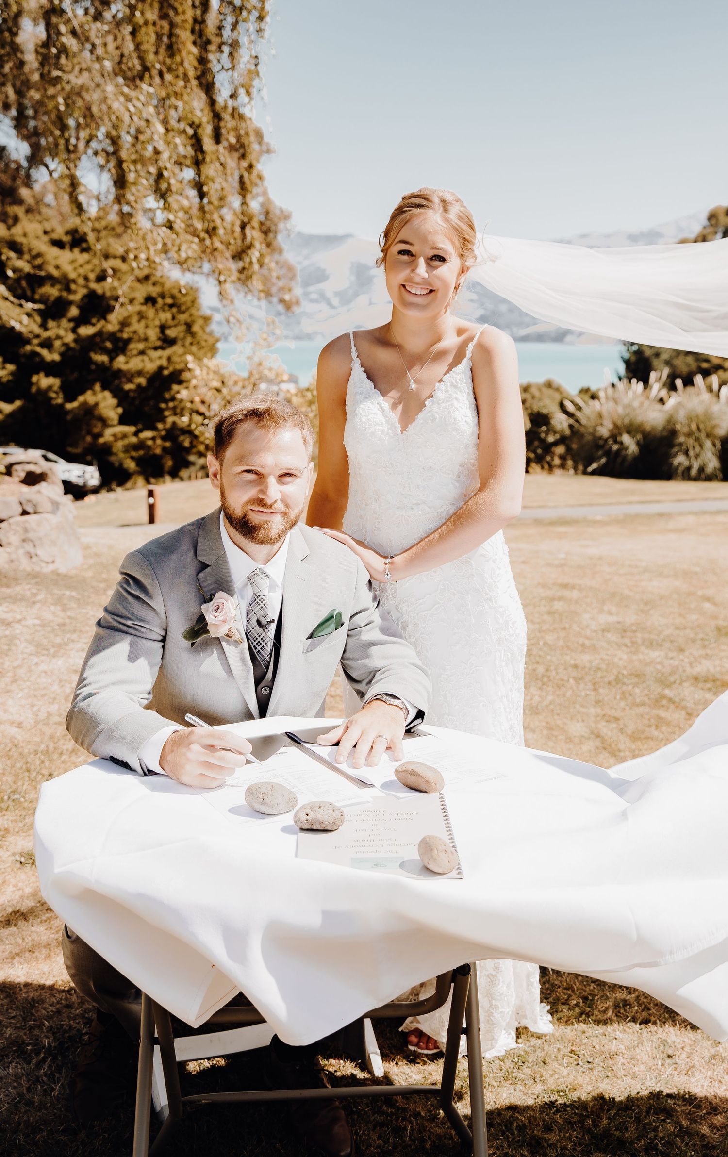 Couple poses at signing table during outdoor wedding with mountain vista behind them.