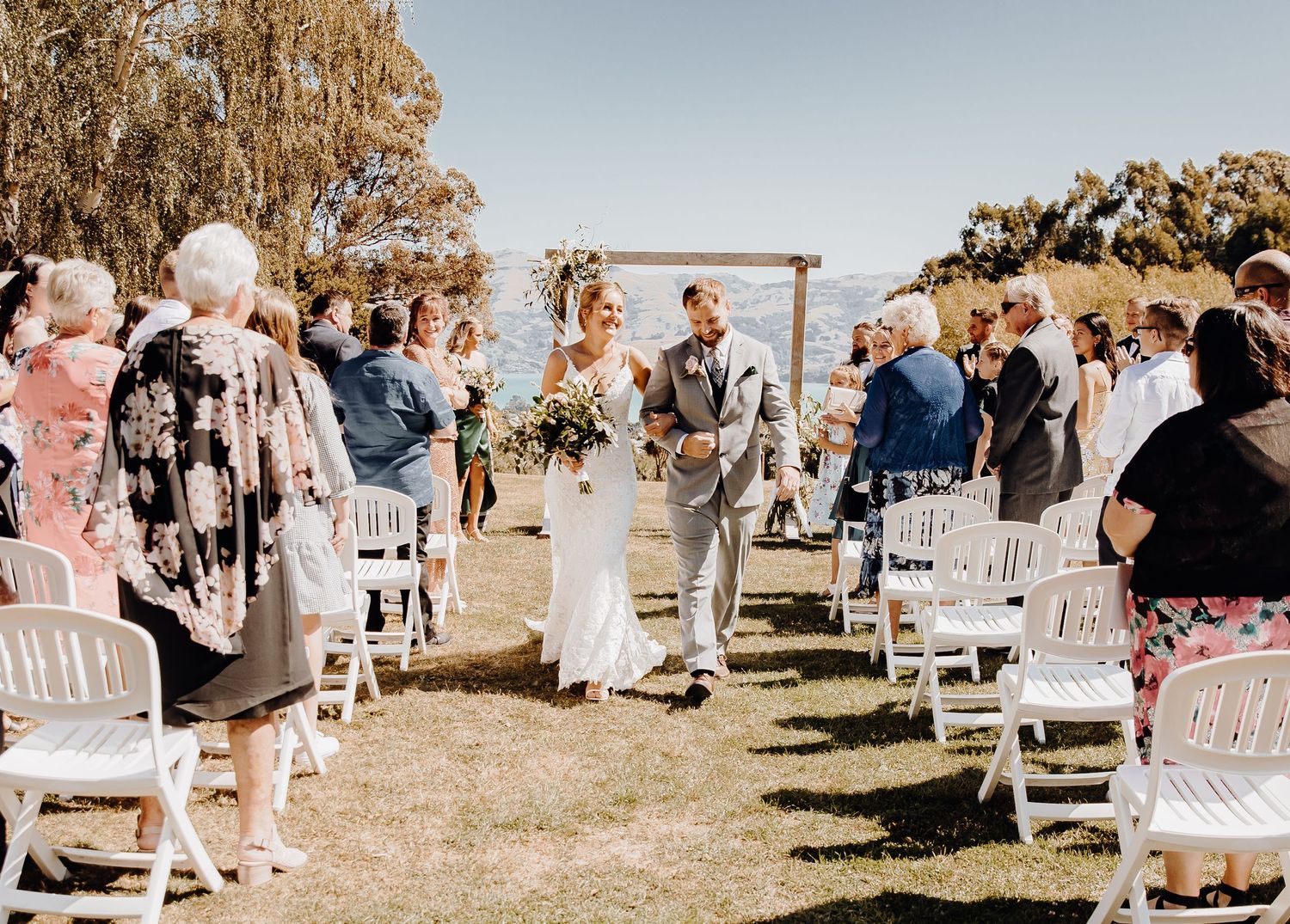 Wedding ceremony recessional on grassy hill with white chairs and mountain backdrop.