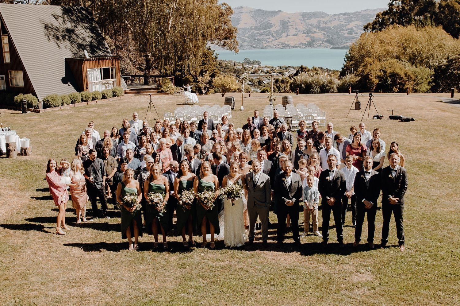 Large wedding group photo of guests and wedding party on hillside overlooking lake.