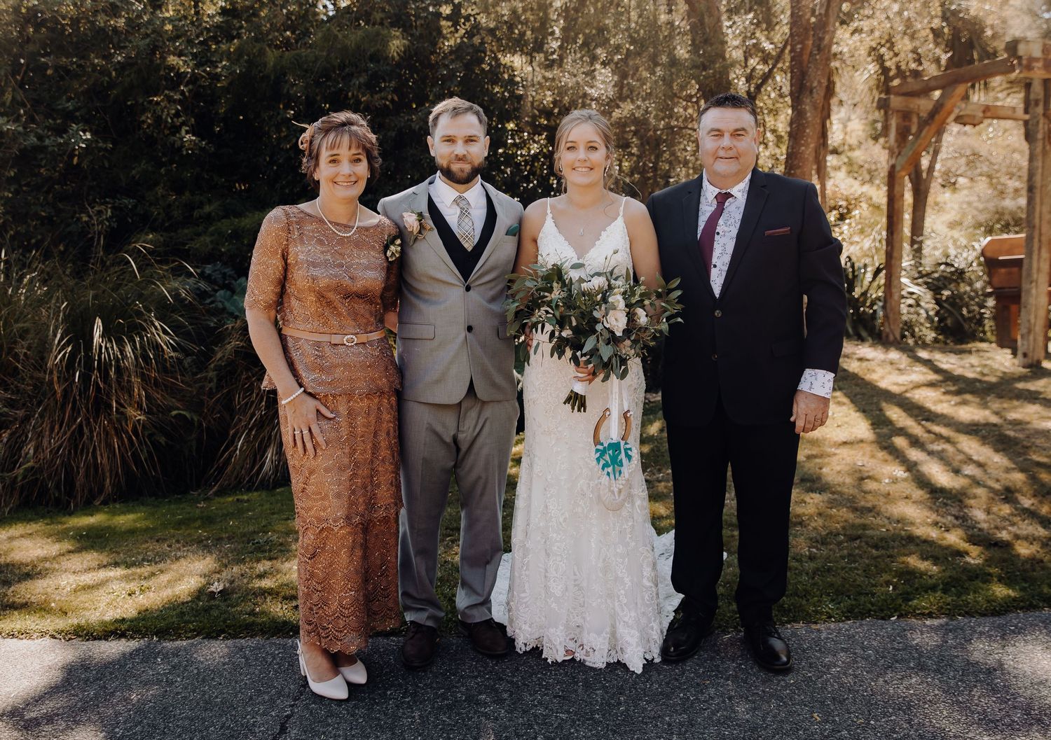 Four wedding guests pose together outdoors in formal attire against a sunlit natural backdrop.