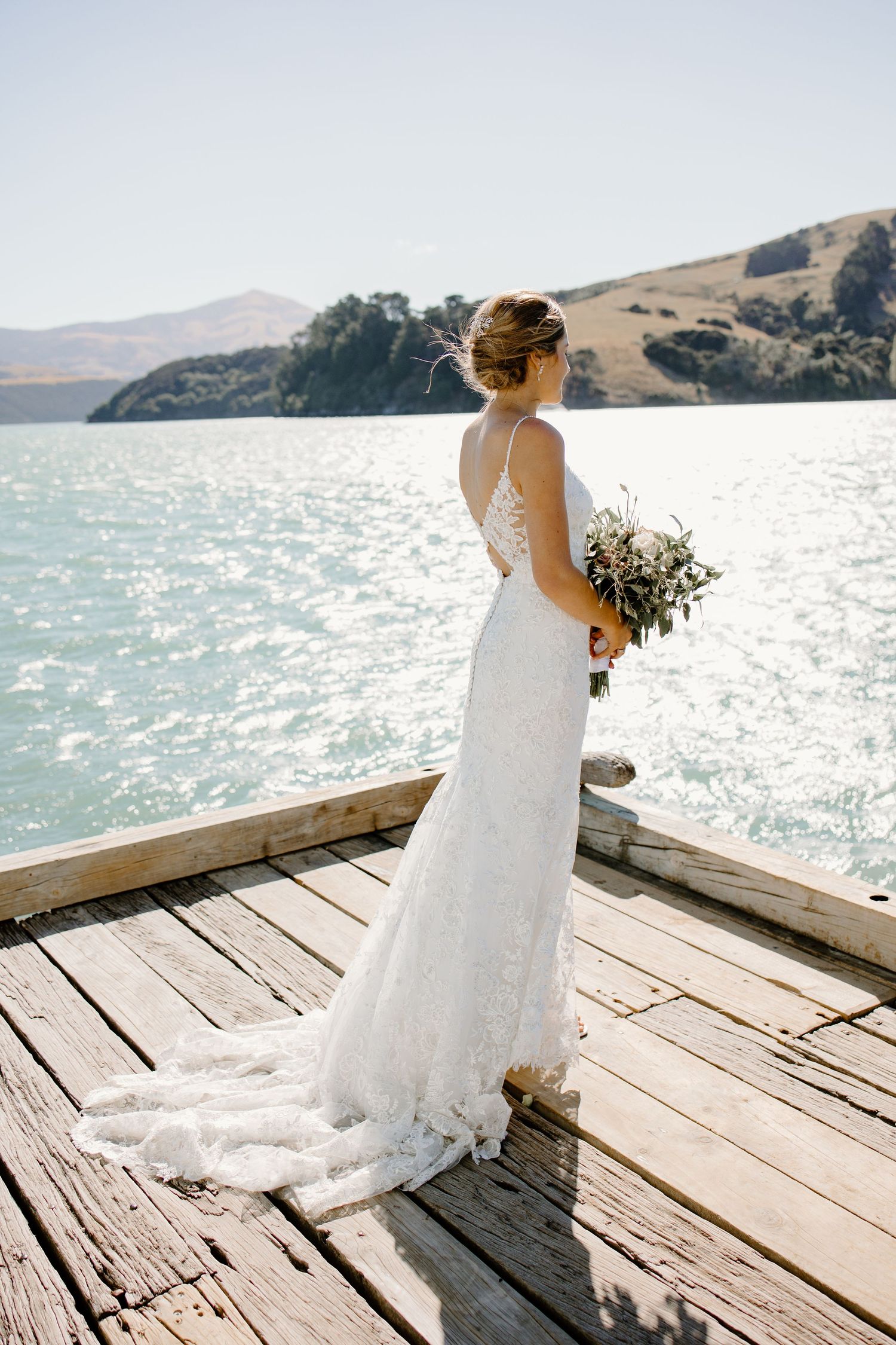Bride in lace dress stands on wooden dock overlooking scenic water view.