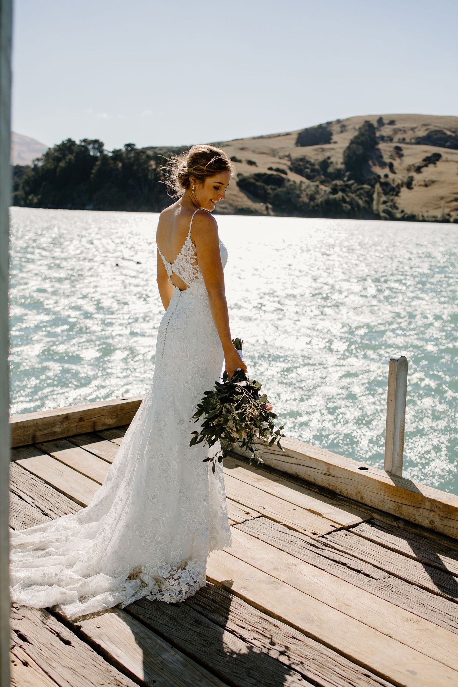 Back view of bride in lace gown standing on dock with water and hills in background.