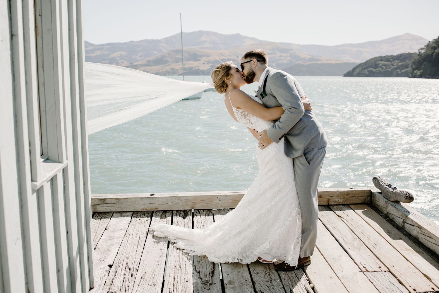 A romantic wedding couple shares a kiss on a wooden dock overlooking a scenic mountain lake.