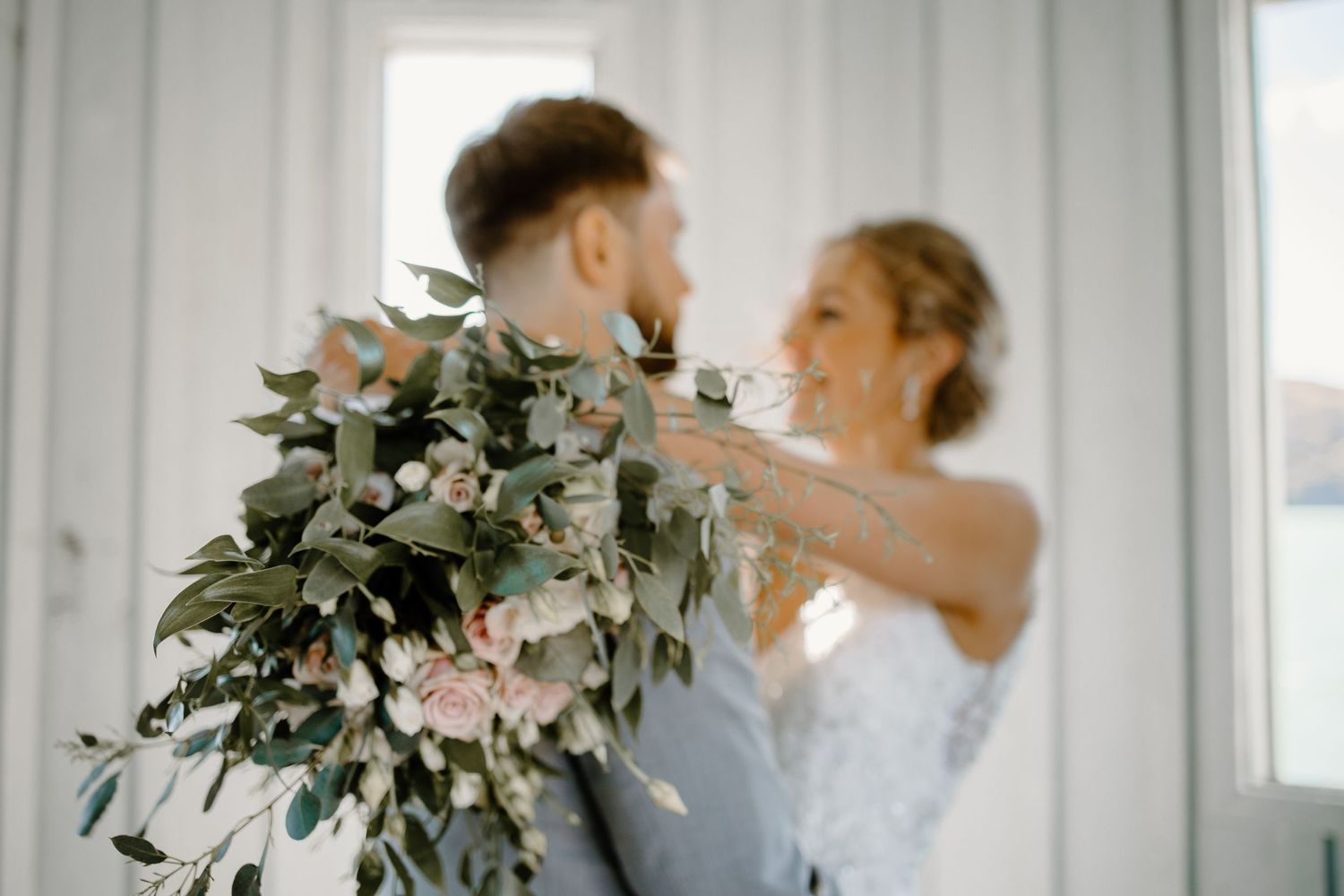 A wedding couple shares a joyful moment with a lush eucalyptus and rose bouquet indoors.