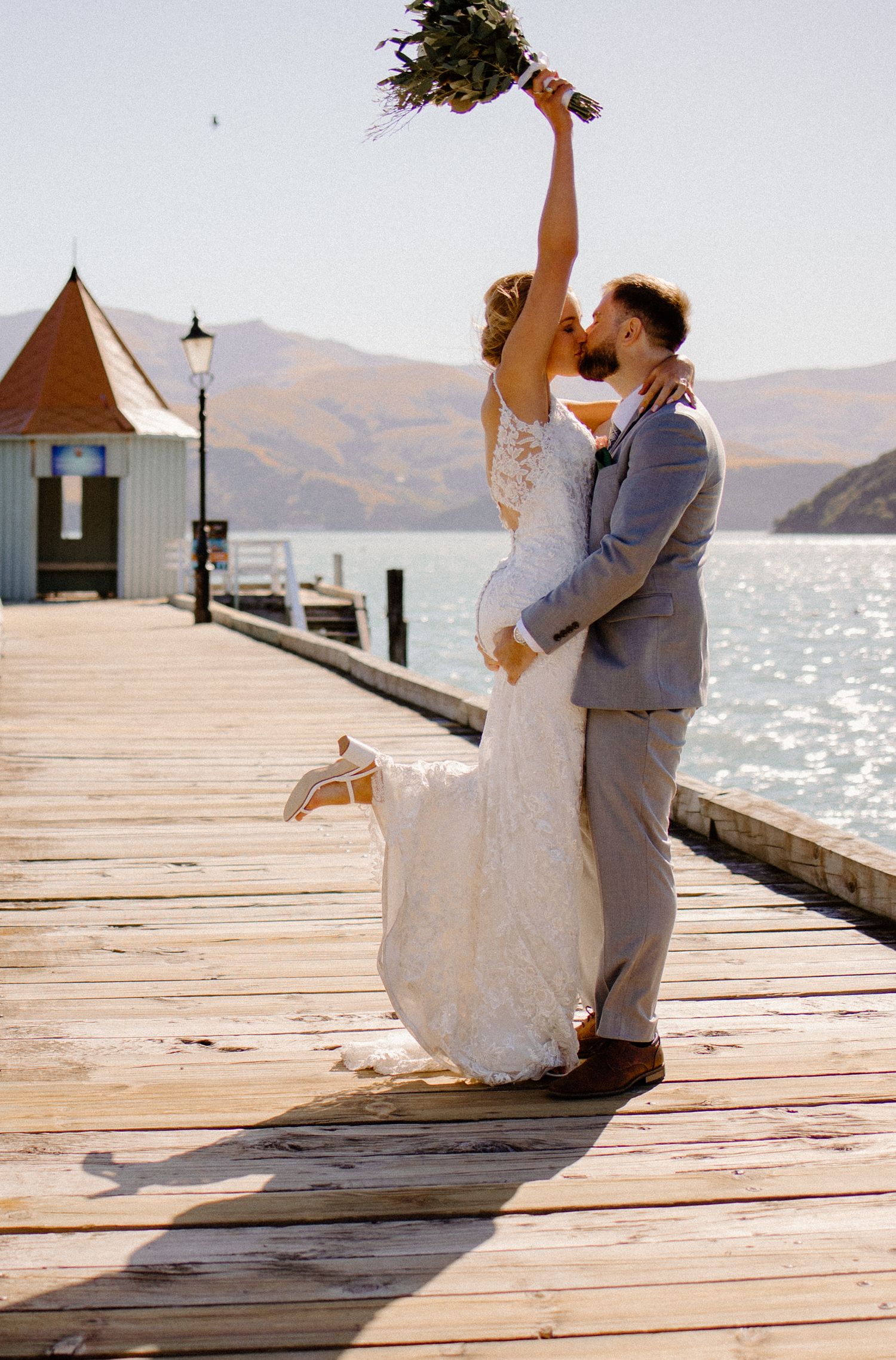 A bride holds her bouquet high while embracing on a scenic waterfront dock.