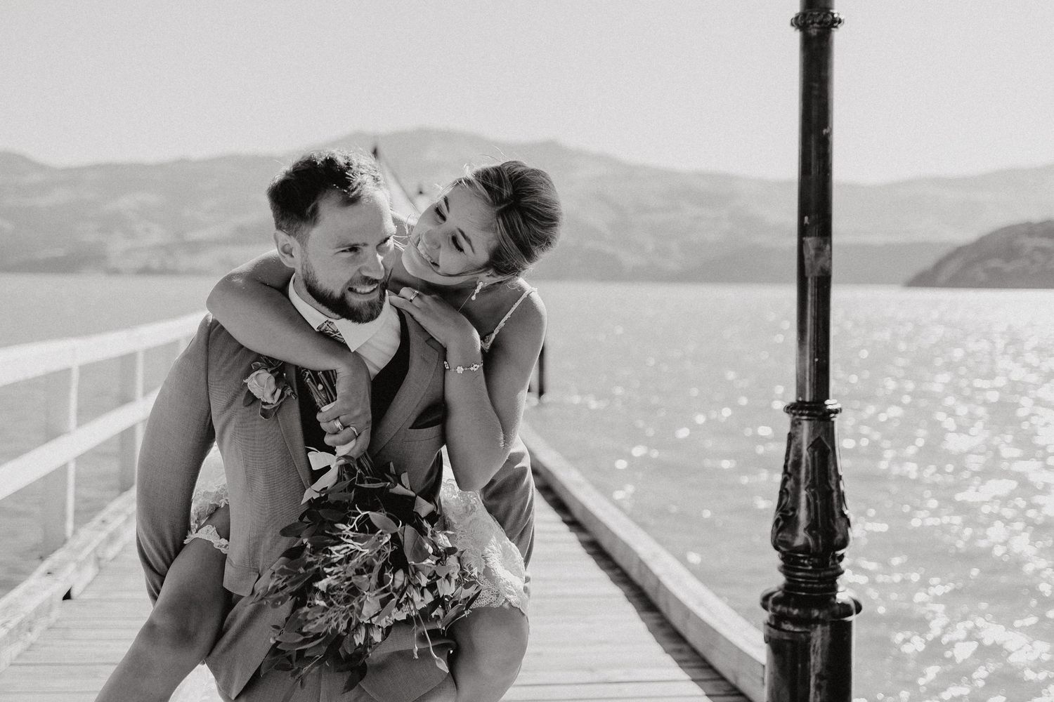 A playful black and white wedding photo captured on a pier with mountains in the background.