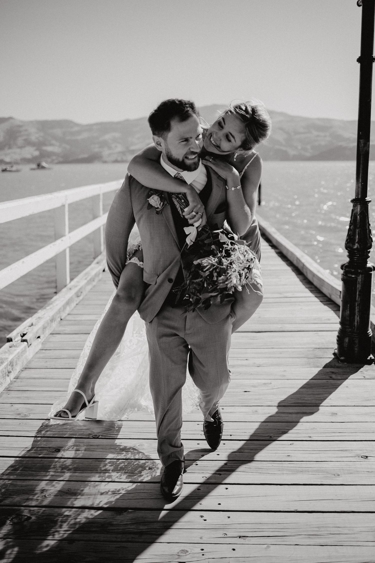 A black and white photo of a groom carrying his bride on a wooden pier.