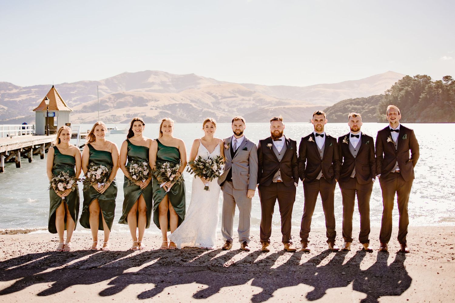 Wedding party in green dresses and dark suits poses on a sunny dock by the water.