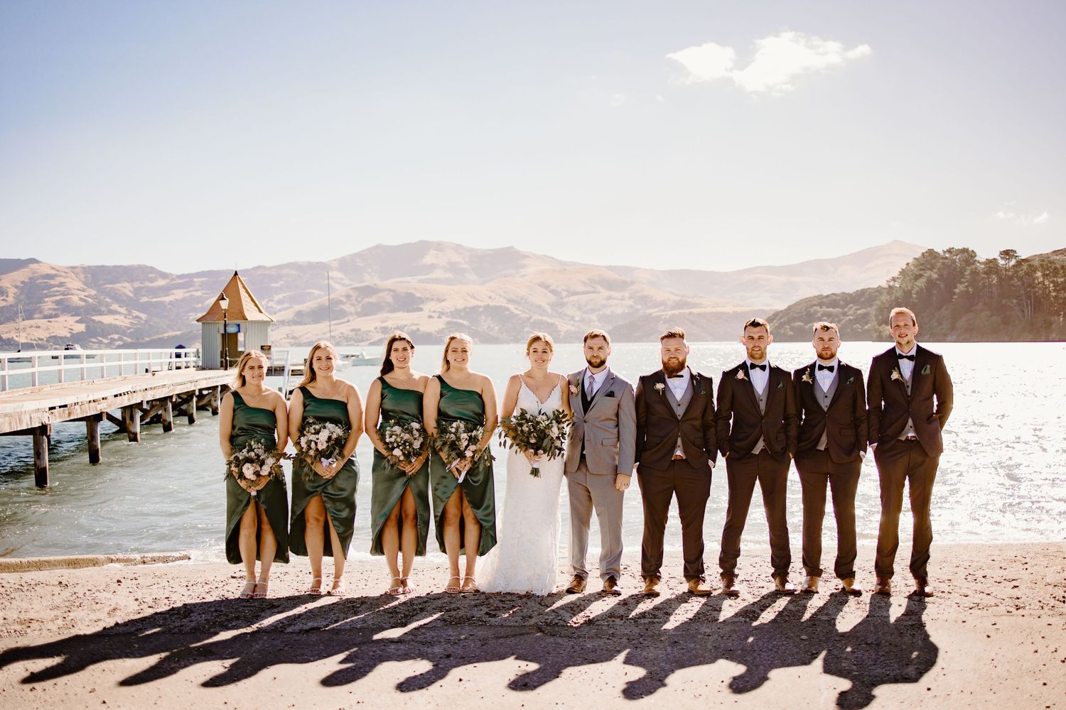 Wedding party lined up on a sunny pier with bridesmaids in green and groomsmen in dark suits.