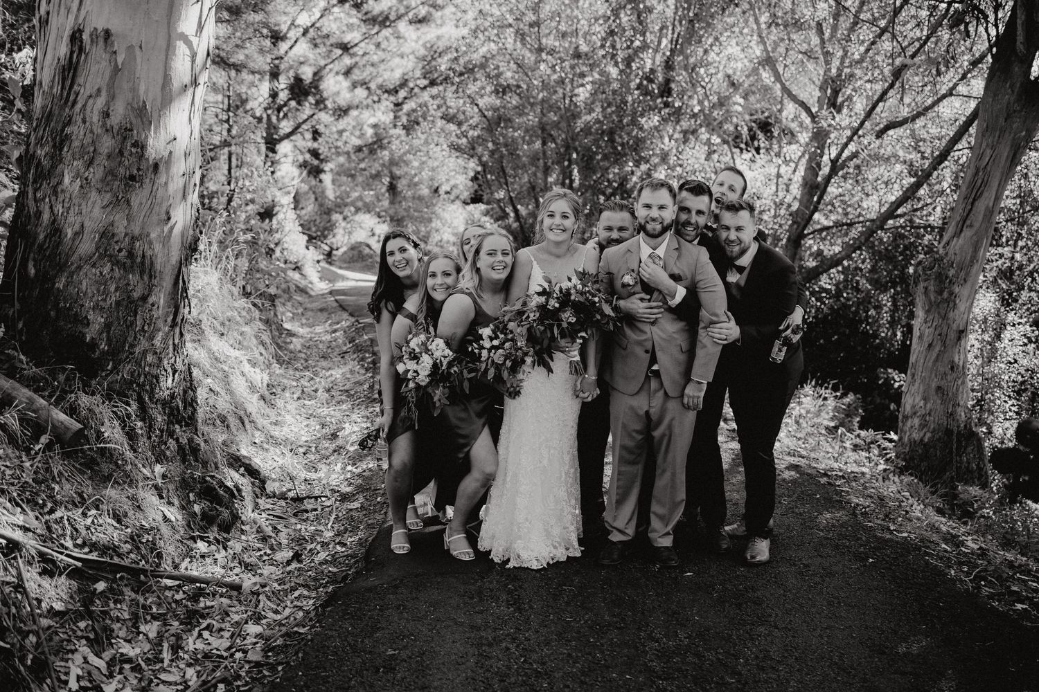 A wedding party poses together on a forest path surrounded by tall trees.