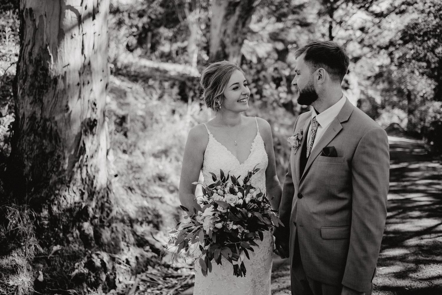 A couple shares an intimate moment on a wooded path as they hold a wedding bouquet.