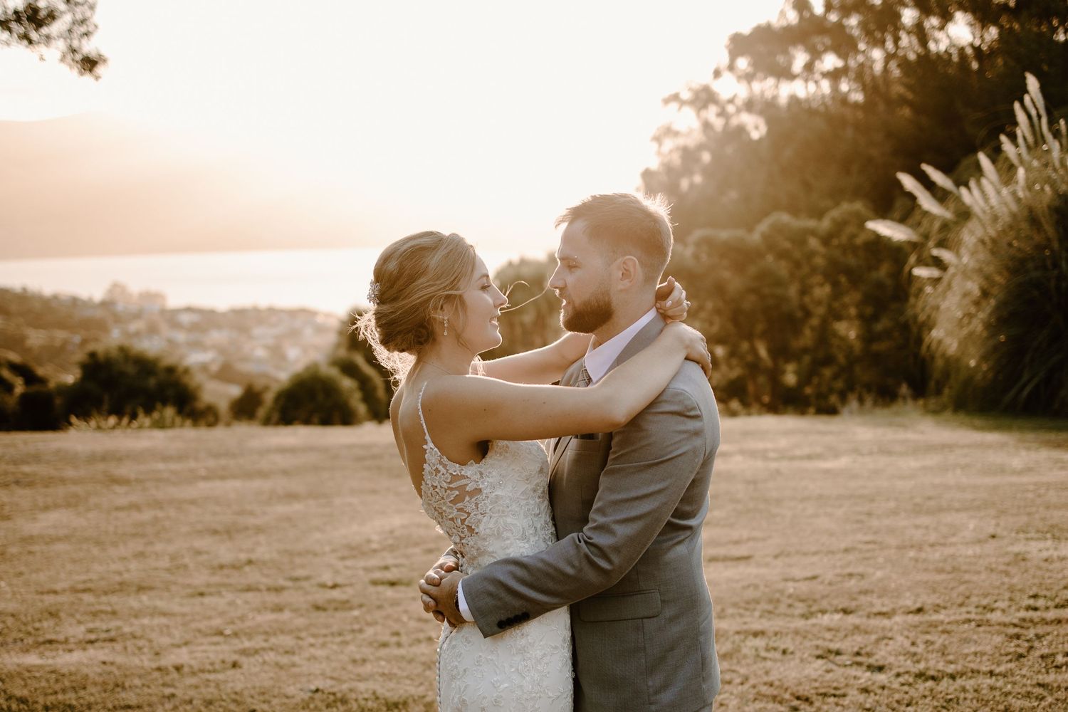 A romantic sunset wedding portrait shows a couple embracing in a lace gown and gray suit on a hilltop overlook.