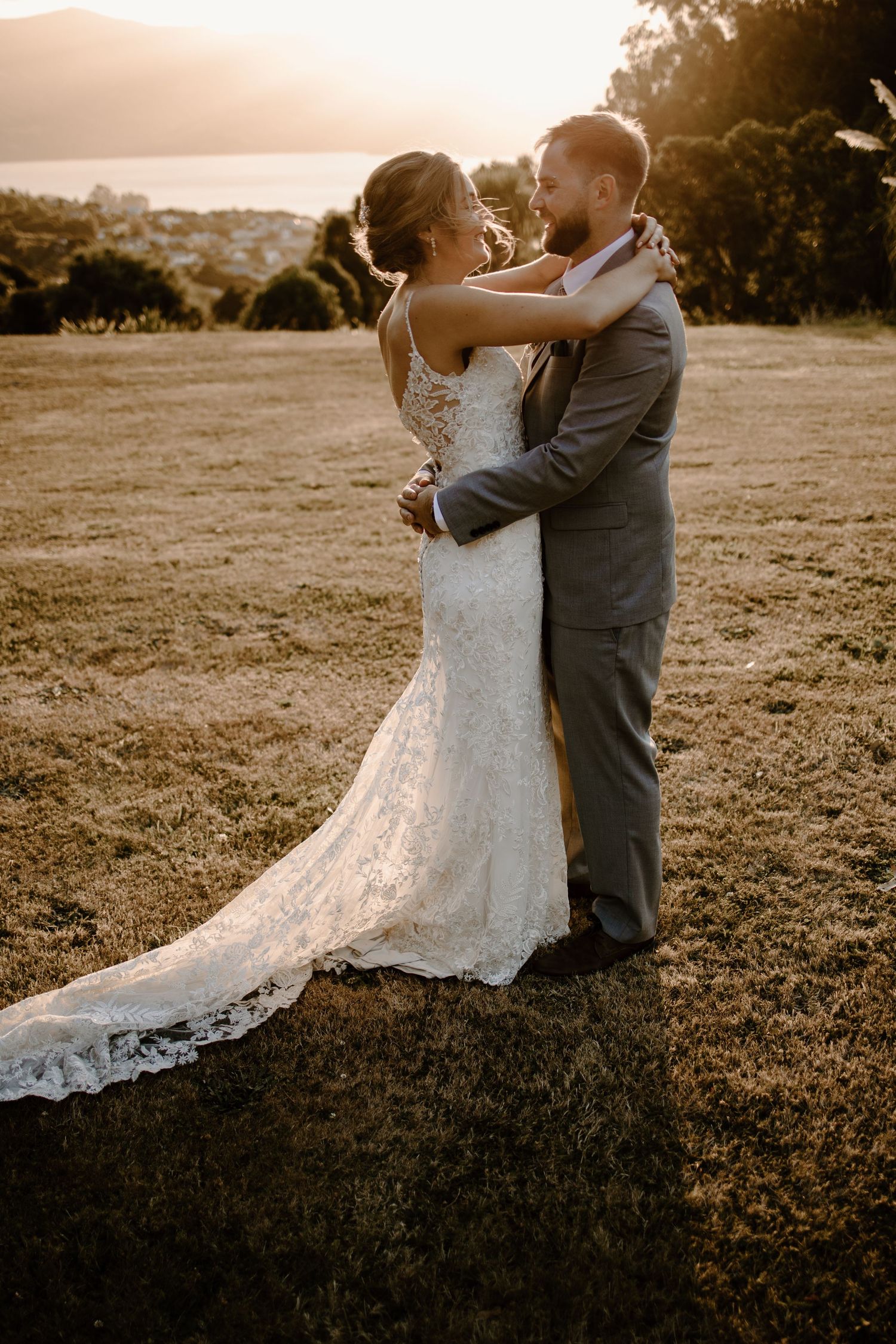 Newlyweds share an intimate moment at golden hour with the bride wearing a flowing lace gown and groom in gray.