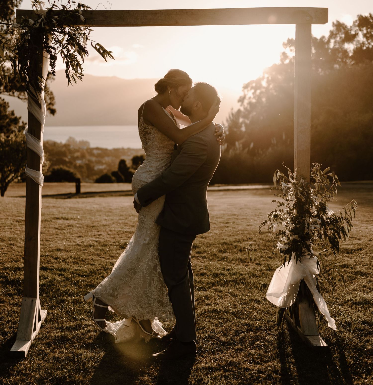 A couple shares their first kiss under a wooden wedding arch at sunset with mountains in the background.