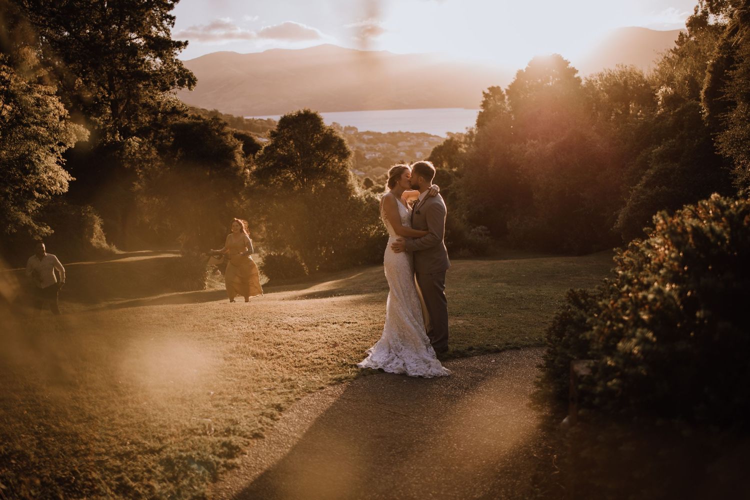 Newlyweds embrace during golden hour on a hillside path with dramatic sunlight streaming through trees.