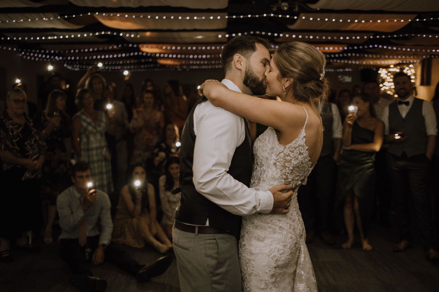 A romantic first dance moment unfolds under string lights at an indoor wedding reception.