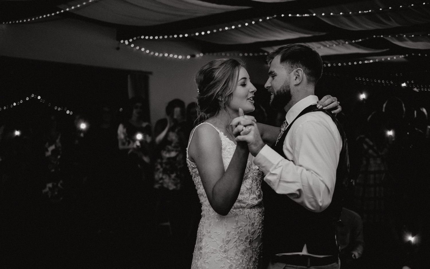 A black and white reception photo captures a joyful dancing moment under string lights.