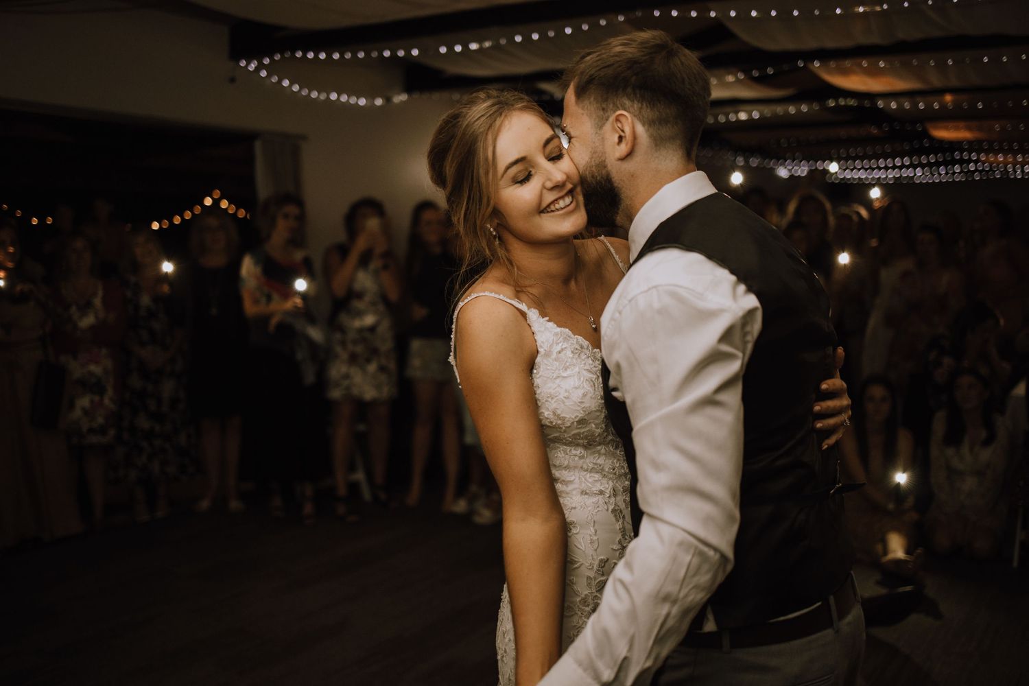 Romantic first dance moment between newlyweds at dimly lit wedding reception with string lights overhead.