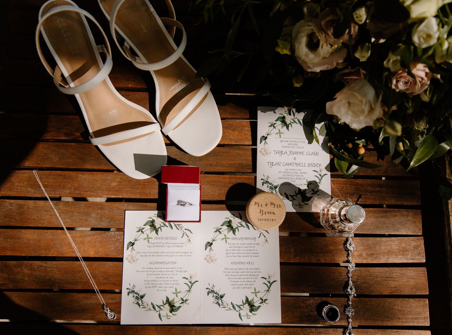 Wedding details flatlay with white sandals, floral invitations and ring box on wooden surface.
