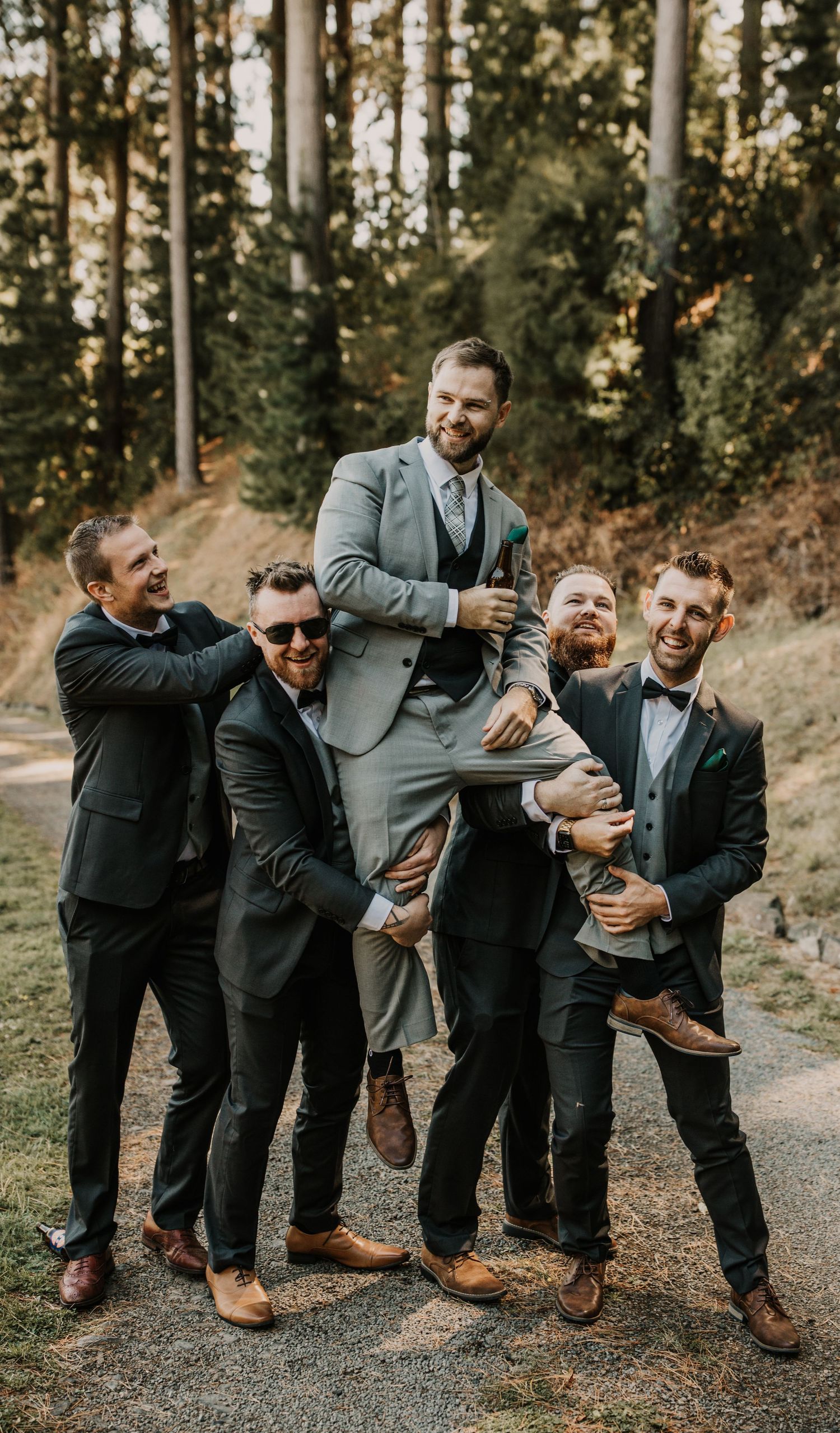 Groomsmen in dark suits playfully lift and carry the groom in a light gray suit on a wooded outdoor path.