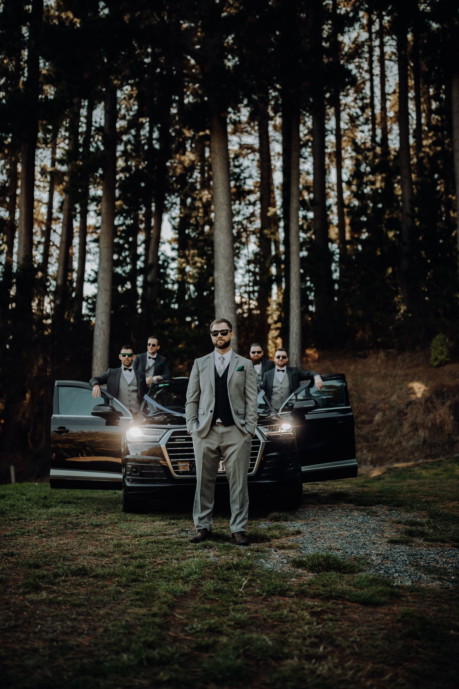 Wedding party poses with luxury car in moody forest setting at dusk.