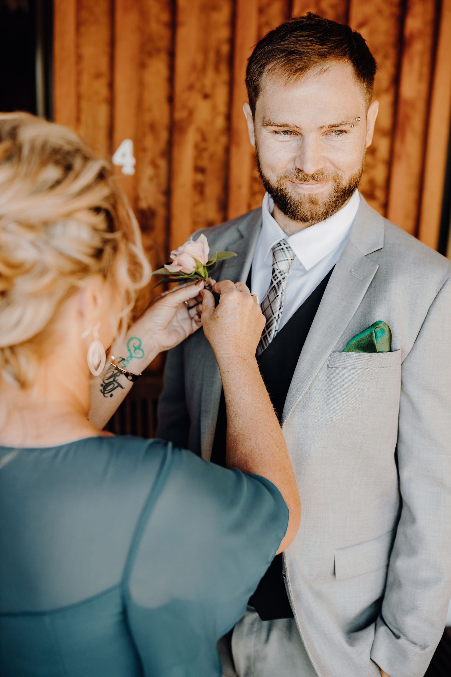 Person in teal dress pins boutonniere on light gray suit during wedding preparation.