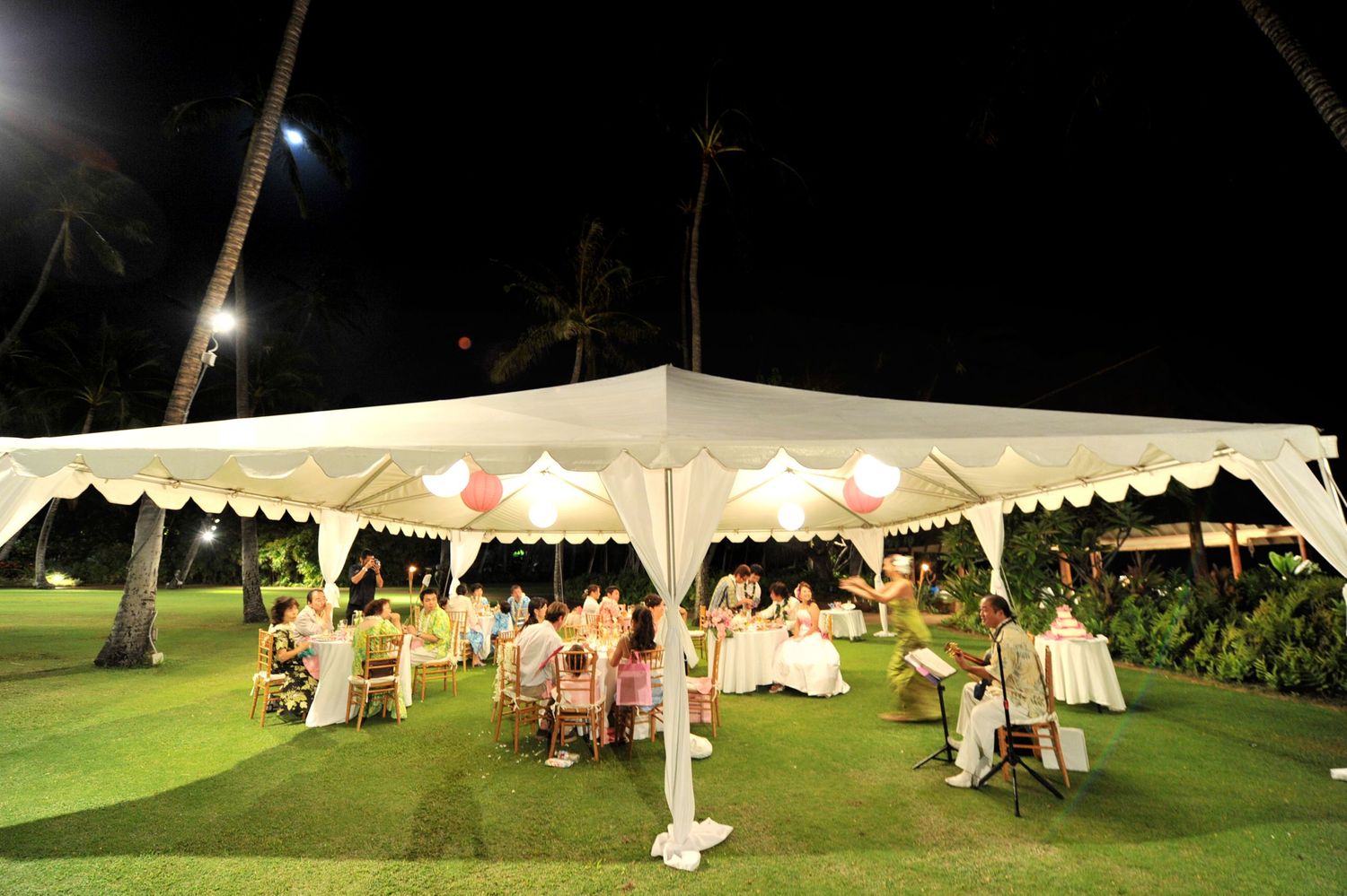 Festive outdoor wedding reception under a white tent at night with colorful decorations and palm trees.