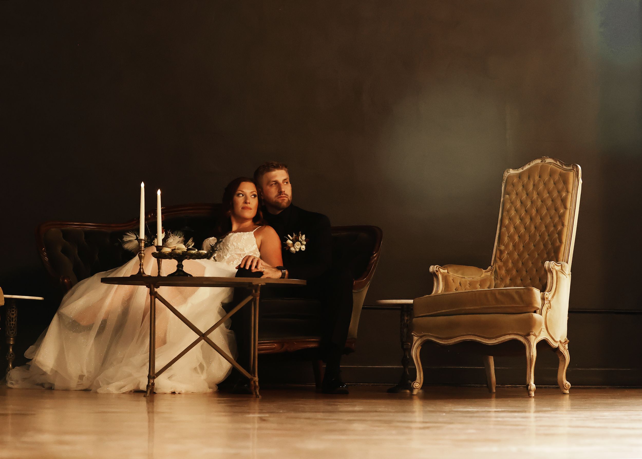 A romantic wedding couple sits at an elegantly set table with candles and vintage furniture in a moody setting.