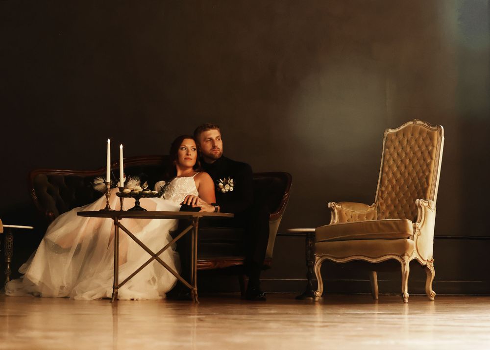 A romantic wedding couple sits at an elegantly set table with candles and vintage furniture in a moody setting.