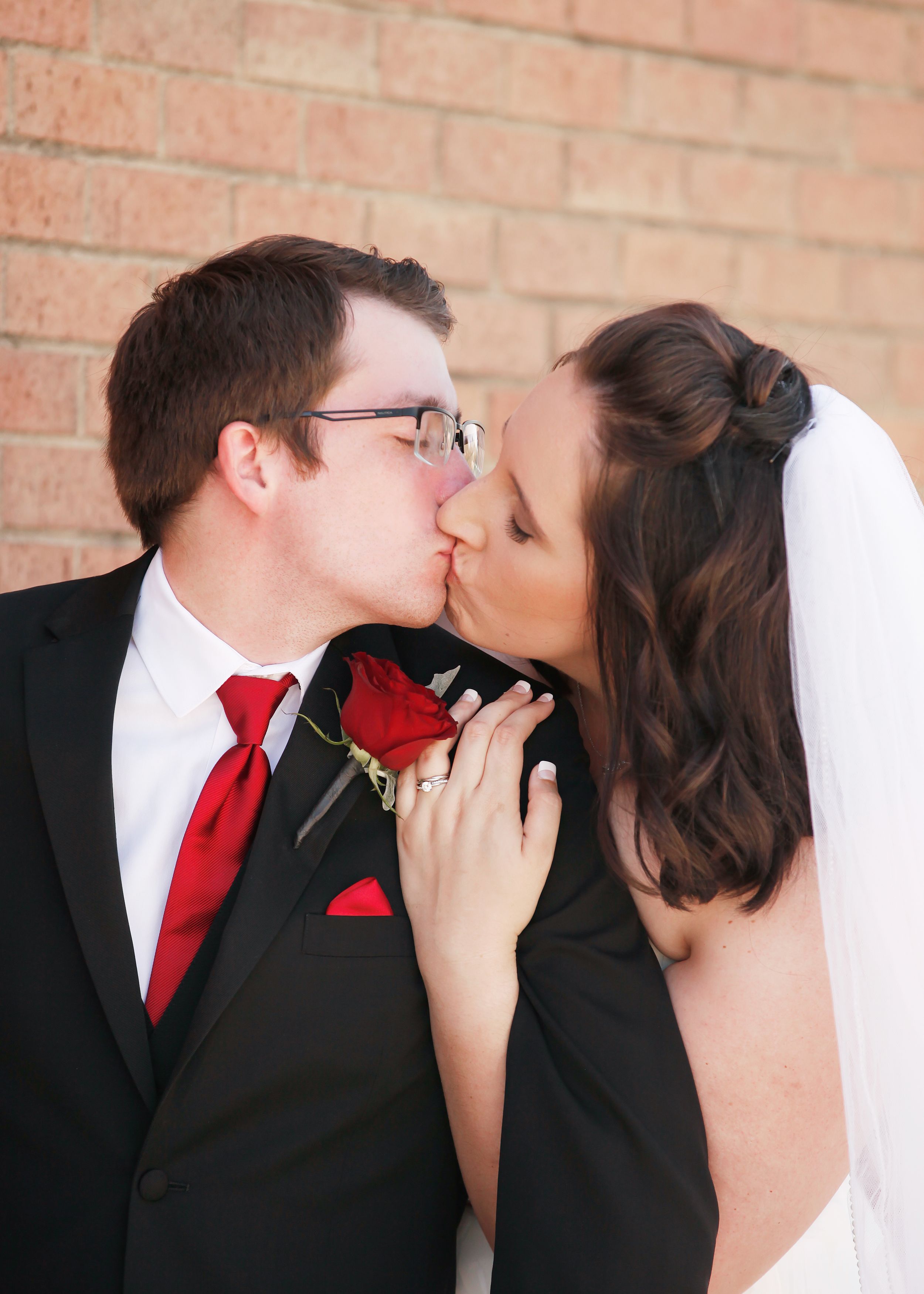 A romantic wedding kiss between newlyweds, with the groom wearing a black suit and red tie against a brick wall.