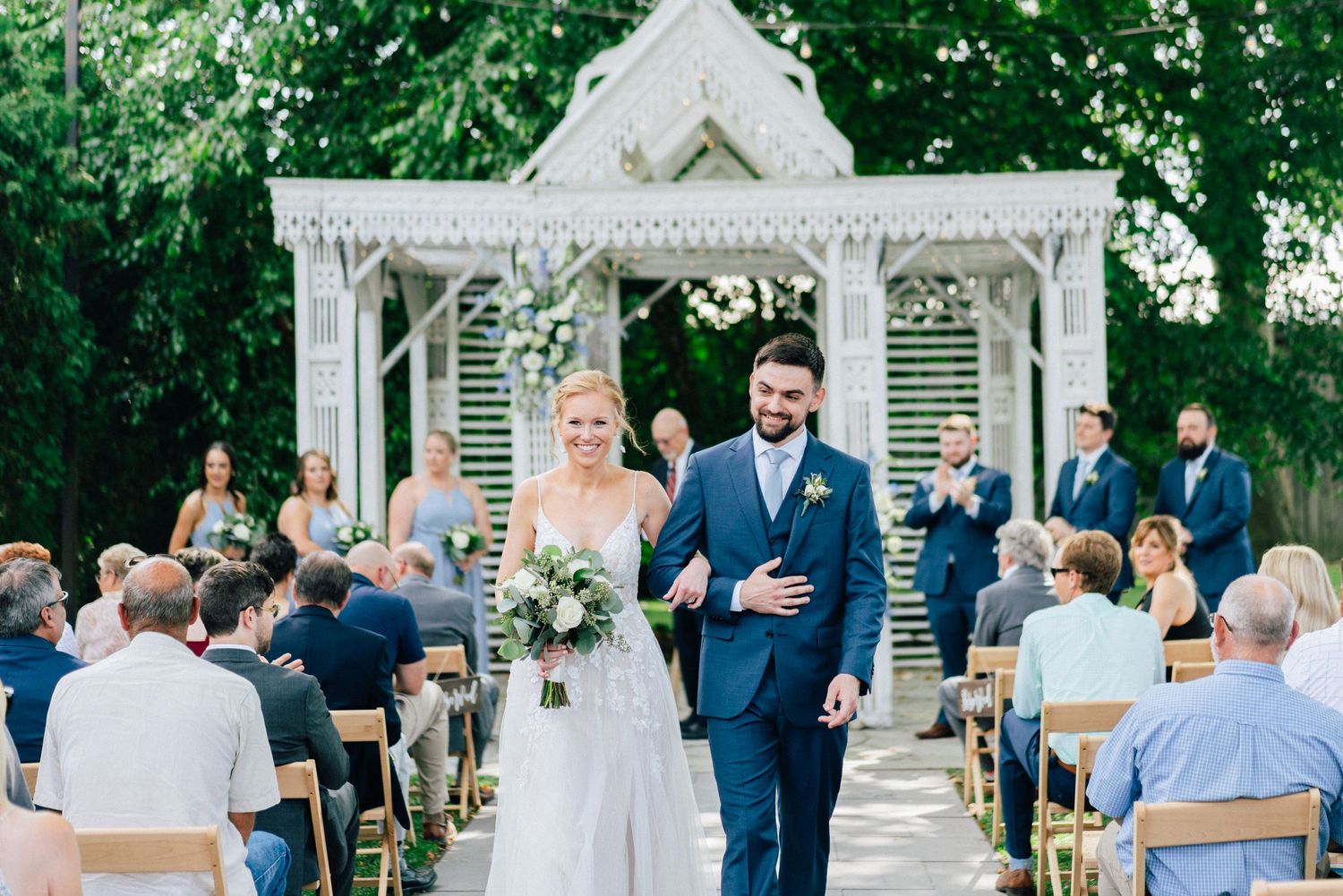 Wedding ceremony at a white gazebo surrounded by guests seated on white chairs in a garden setting.
